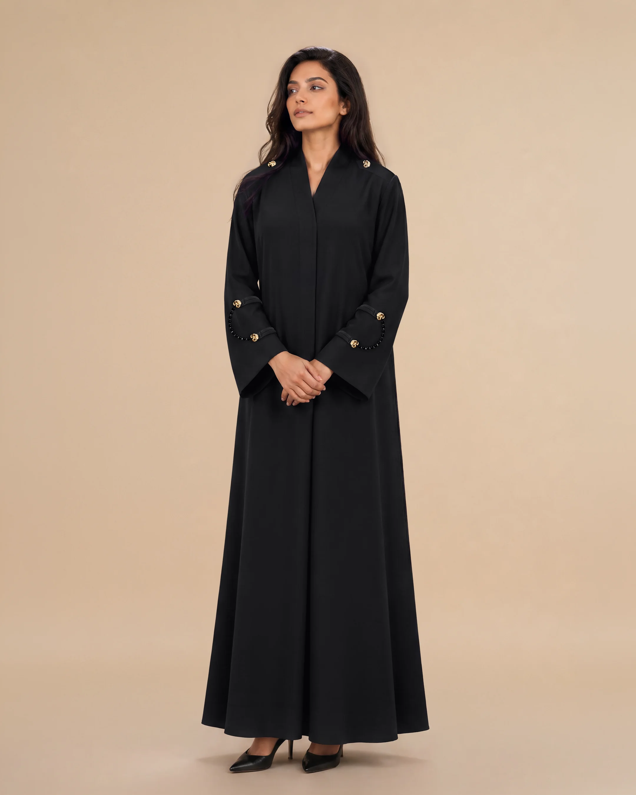 Bint Saeed Marylebone Abaya in Deep Black, front view. Graceful A-line designer abaya with removable genuine natural Onyx Strands on wide cuffs, gold-plated hematite spacer beads, signature gold-tone Knotted Line strand details, hidden side seam pockets, and softly textured crepe — the abaya with natural stone jewellery for your wardrobe. Luxury designer abaya with natural stone jewellery by Bint Saeed Abu Dhabi, United Arab Emirates — graceful A-line abaya with removable Onyx Strands and interchangeable Signature Strands for fashion enthusiasts, fashion editors, curators, cultural heritage audiences, diplomats, gallery openings, and international wardrobes in London, Paris, Riyadh, Doha, and worldwide. Made in Abu Dhabi. Worldwide shipping.