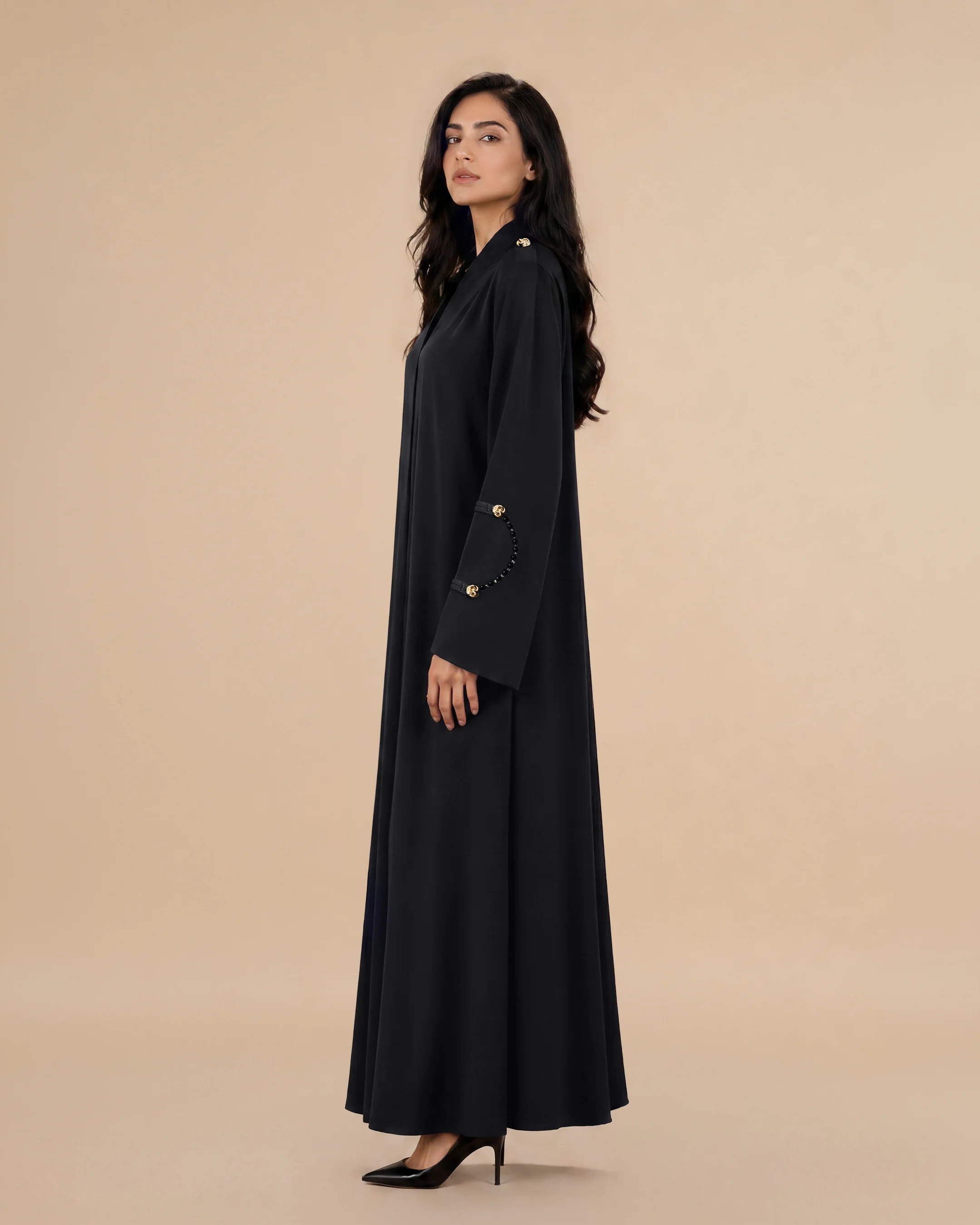 Three-quarter view of the Bint Saeed Marylebone Abaya in Deep Black showcasing graceful A-line drape, removable Onyx Strands with gold-plated hematite beads on wide interchangeable cuffs, and softly textured crepe silhouette. Luxury designer abaya with natural stone jewellery by Bint Saeed Abu Dhabi, United Arab Emirates — graceful A-line abaya with removable Onyx Strands and interchangeable Signature Strands for fashion enthusiasts, fashion editors, curators, cultural heritage audiences, diplomats, gallery openings, and international wardrobes in London, Paris, Riyadh, Doha, and worldwide. Made in Abu Dhabi. Worldwide shipping.